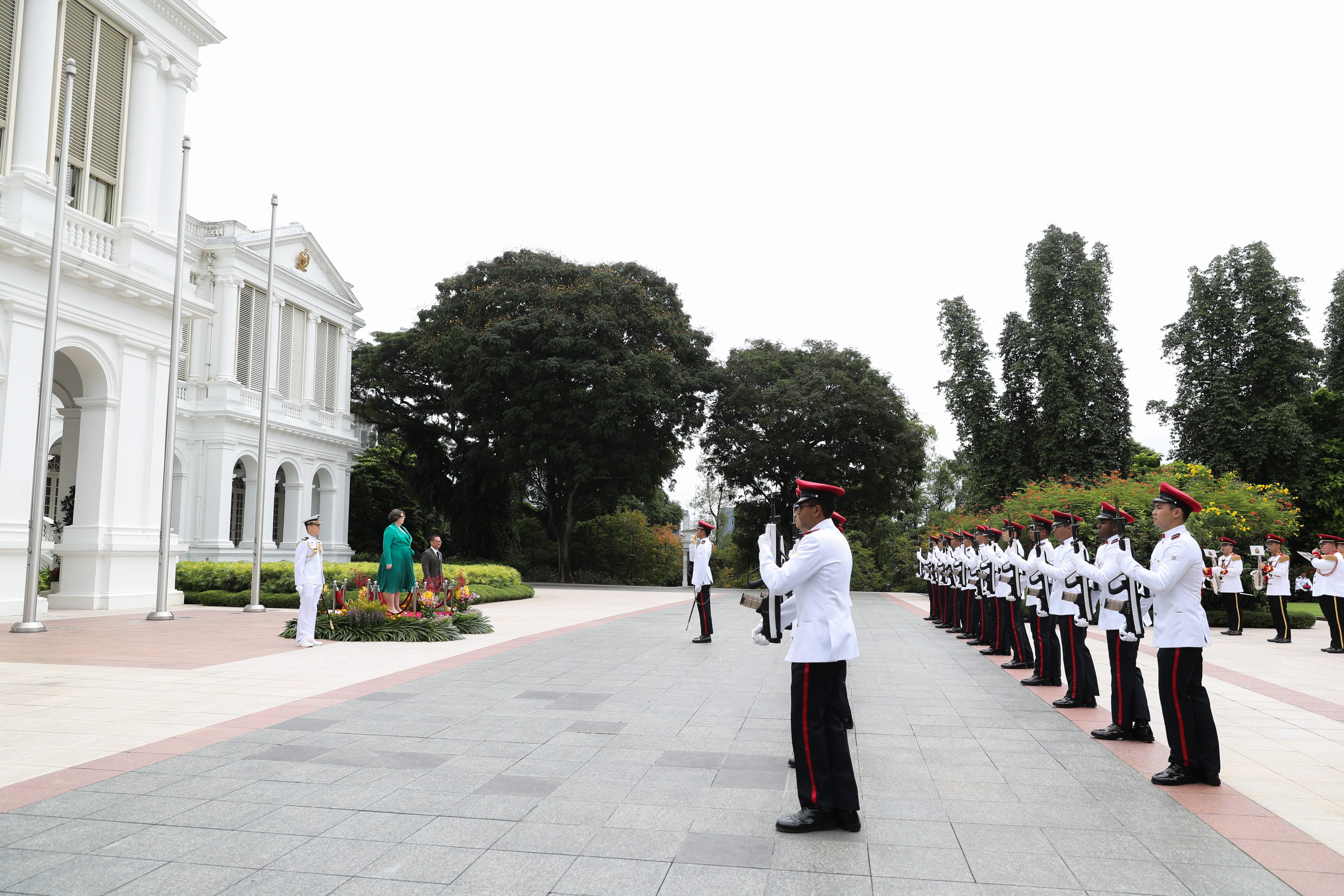 Line of guards in white uniforms with rifles facing two people and a building.
